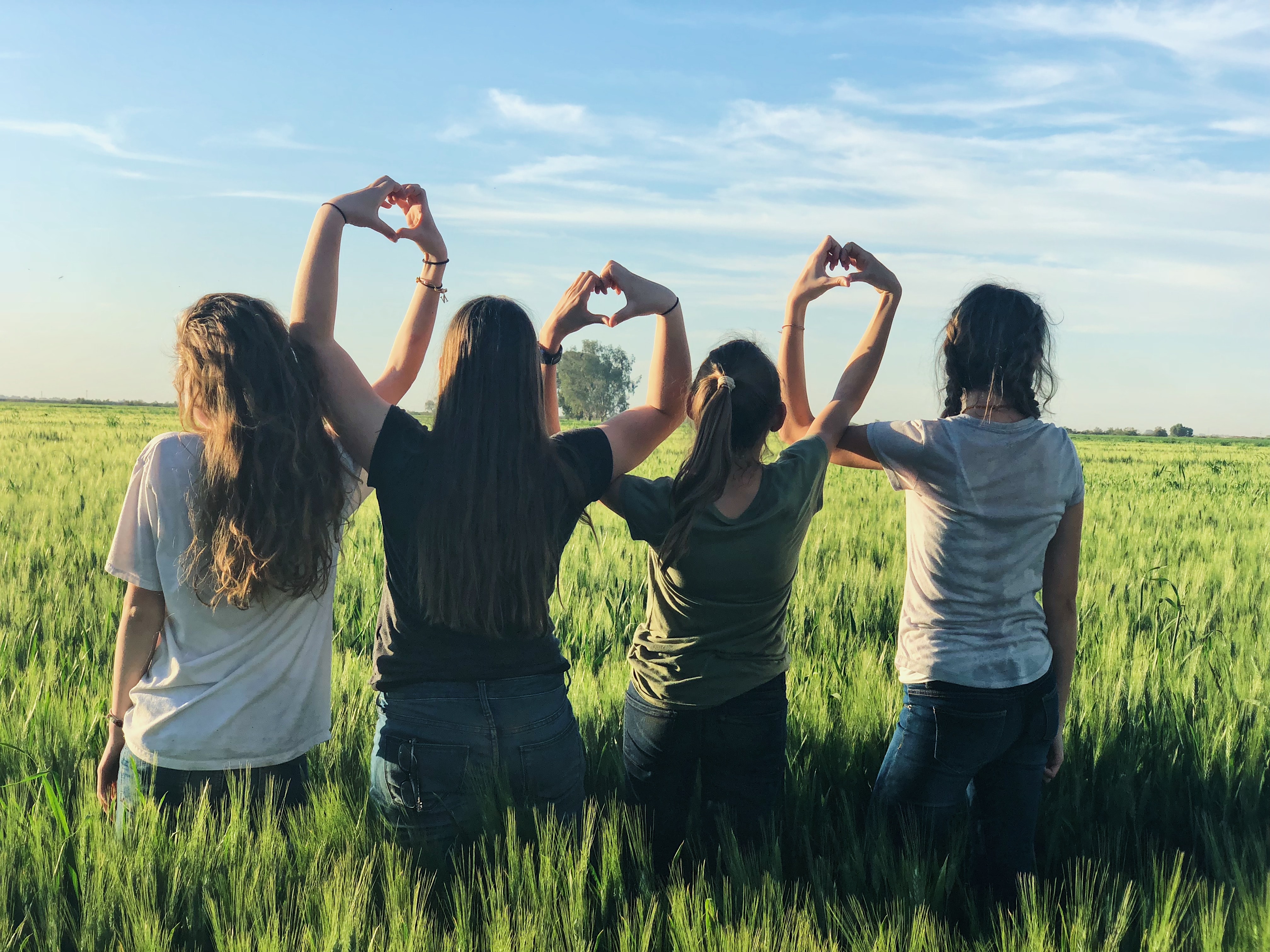 Photo of four young women in a field, backs to the camera, and arms above their head to form hearts with their hands with the person next to them.