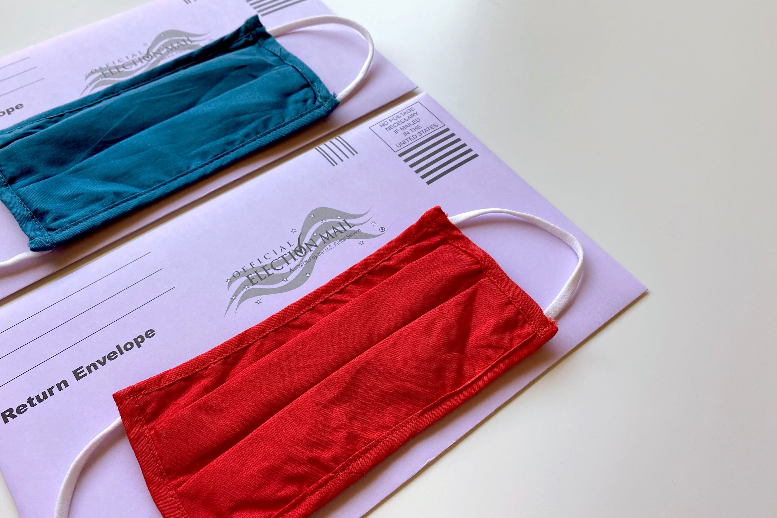 Photo of two Official Election Mail envelopes for mail-in ballots. One envelope has a blue cloth face mask placed on top of it, the other has a red mask.