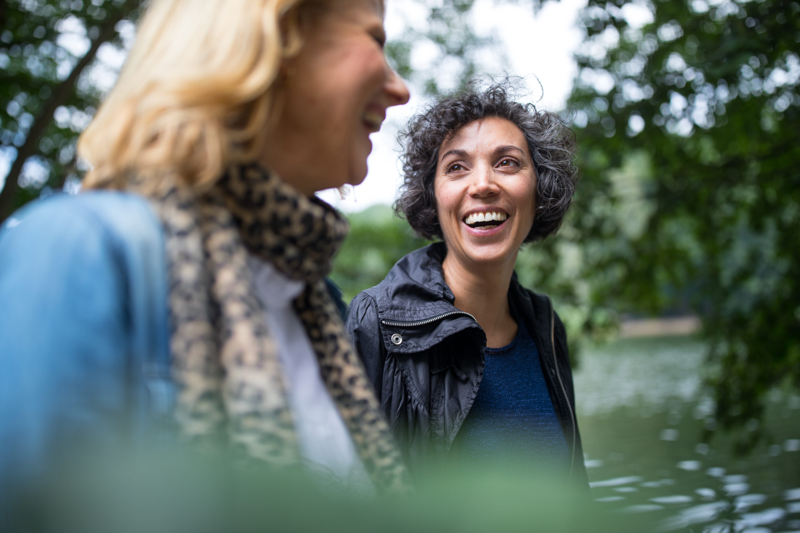 Photo of two smiling women on a walk along a small lake surrounded by trees.