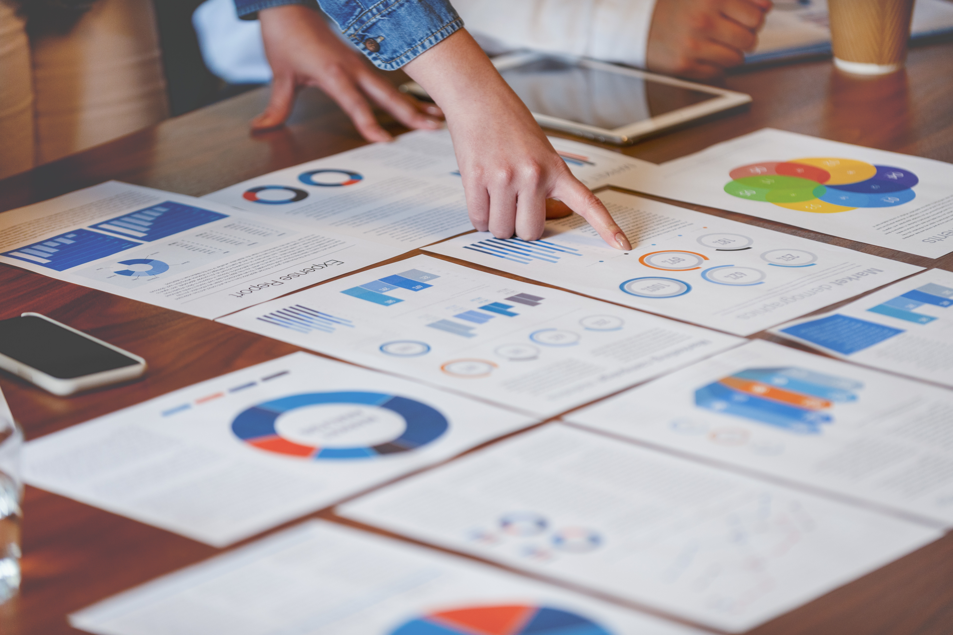 Photo of a conference room table with multiple charts and graphs spread across it and hands pointing at one document.