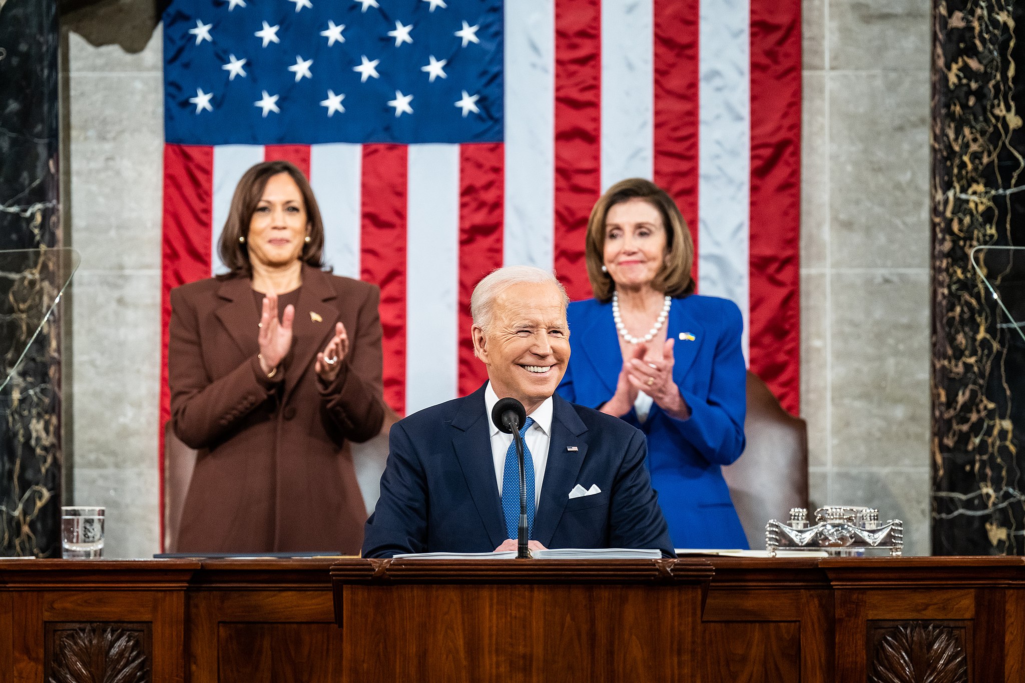 Photo of President Joe Biden delivering State of the Union address with VP Kamala Harris and Speaker Nancy Pelosi standing and applauding behind him.