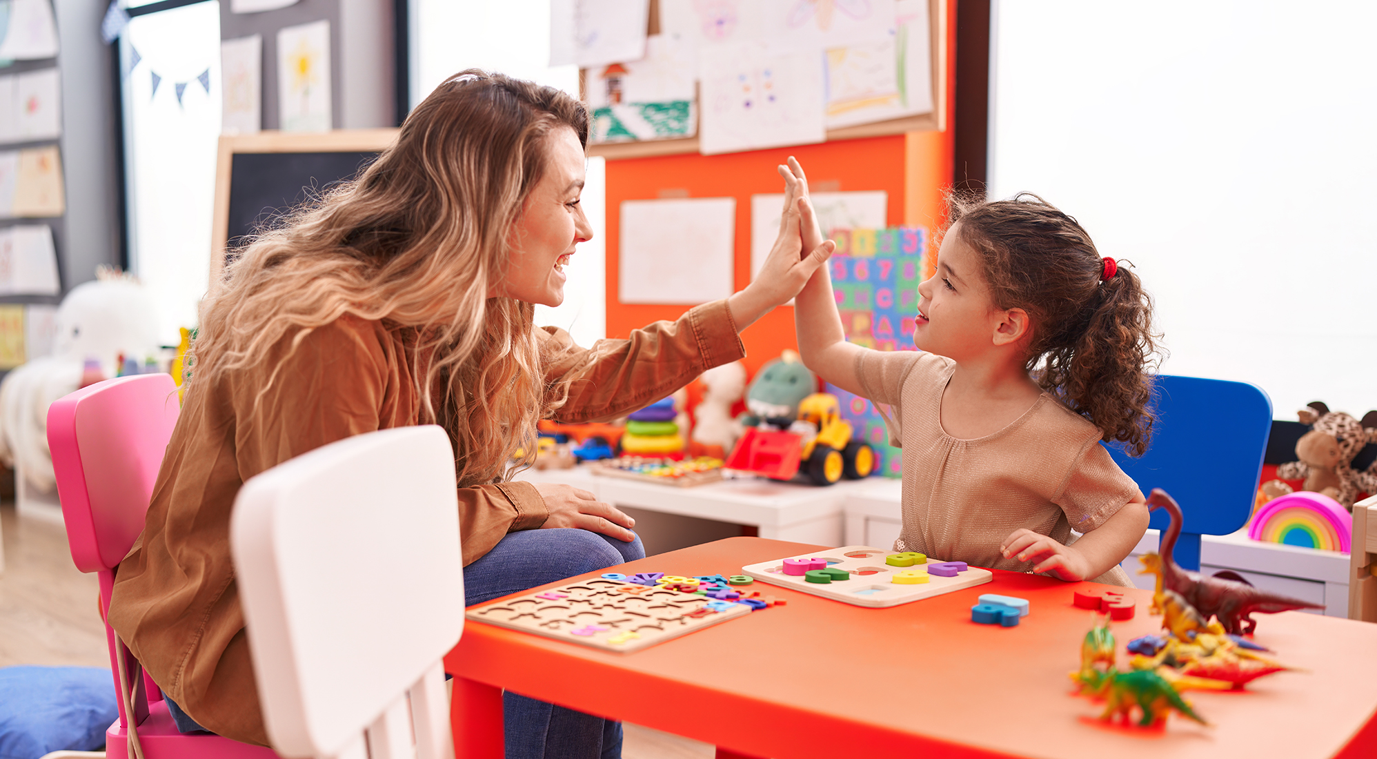 A female daycare worker high fives an adorable child in a daycare setting with lots of toys