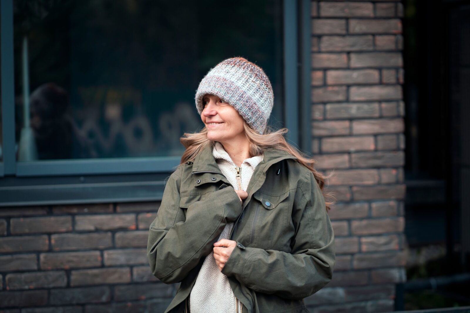 woman outside smiling and looking to the side while wearing a winter coat and hat in front of a brick wall