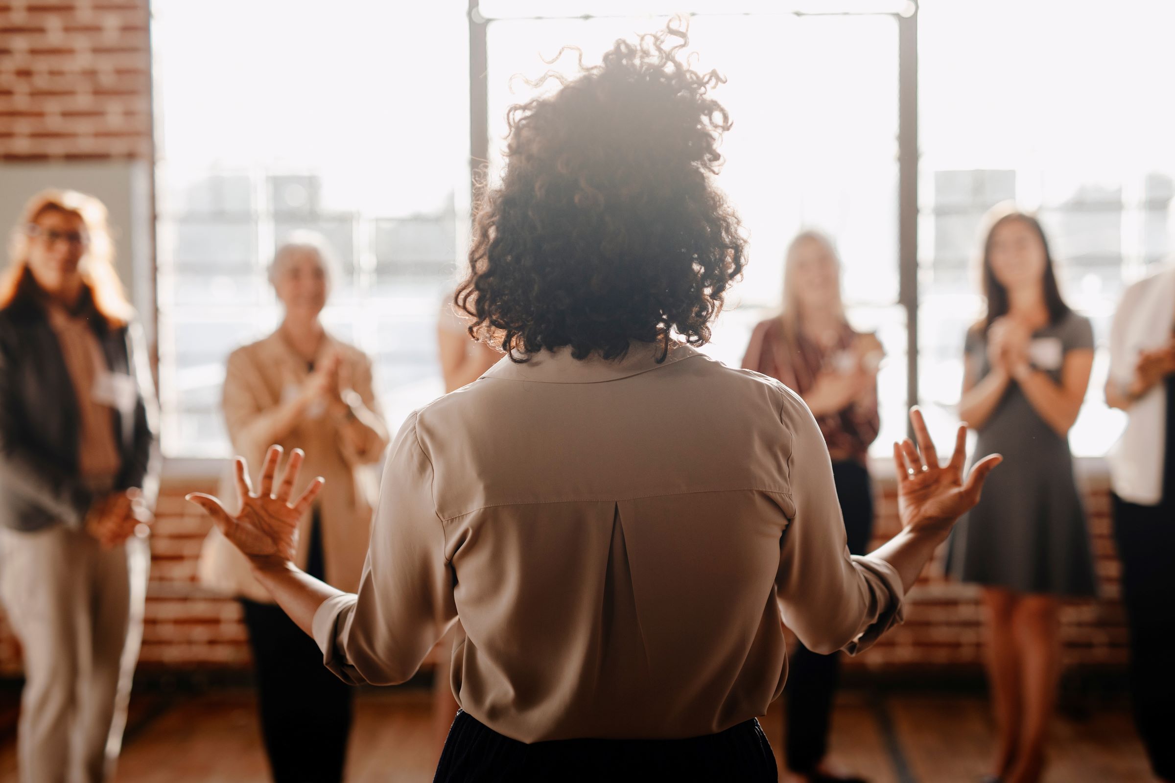 A woman with her back to the camera is addressing an out-of-focus group of women, who are listening attentively.