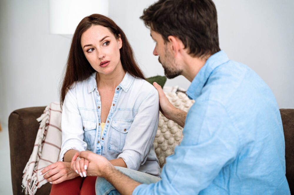 Couple talking on couch looking at each other, man with hand on woman's shoulder
